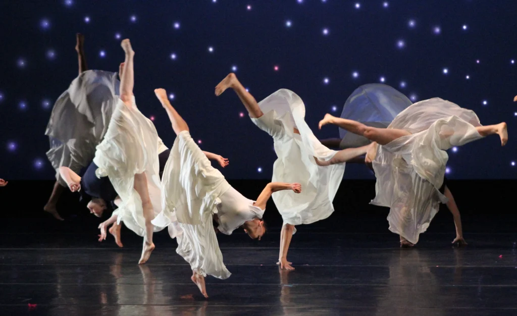 A group of dancers in white dresses are in various stages of flips, bare feet in the air