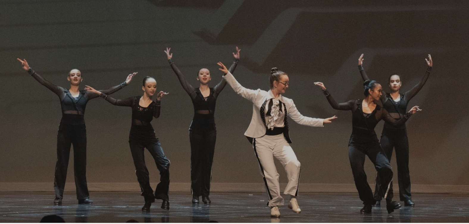 Five tap dancers on a stage, in black pants and sheer black tops surround another tap dancer in a white suit.