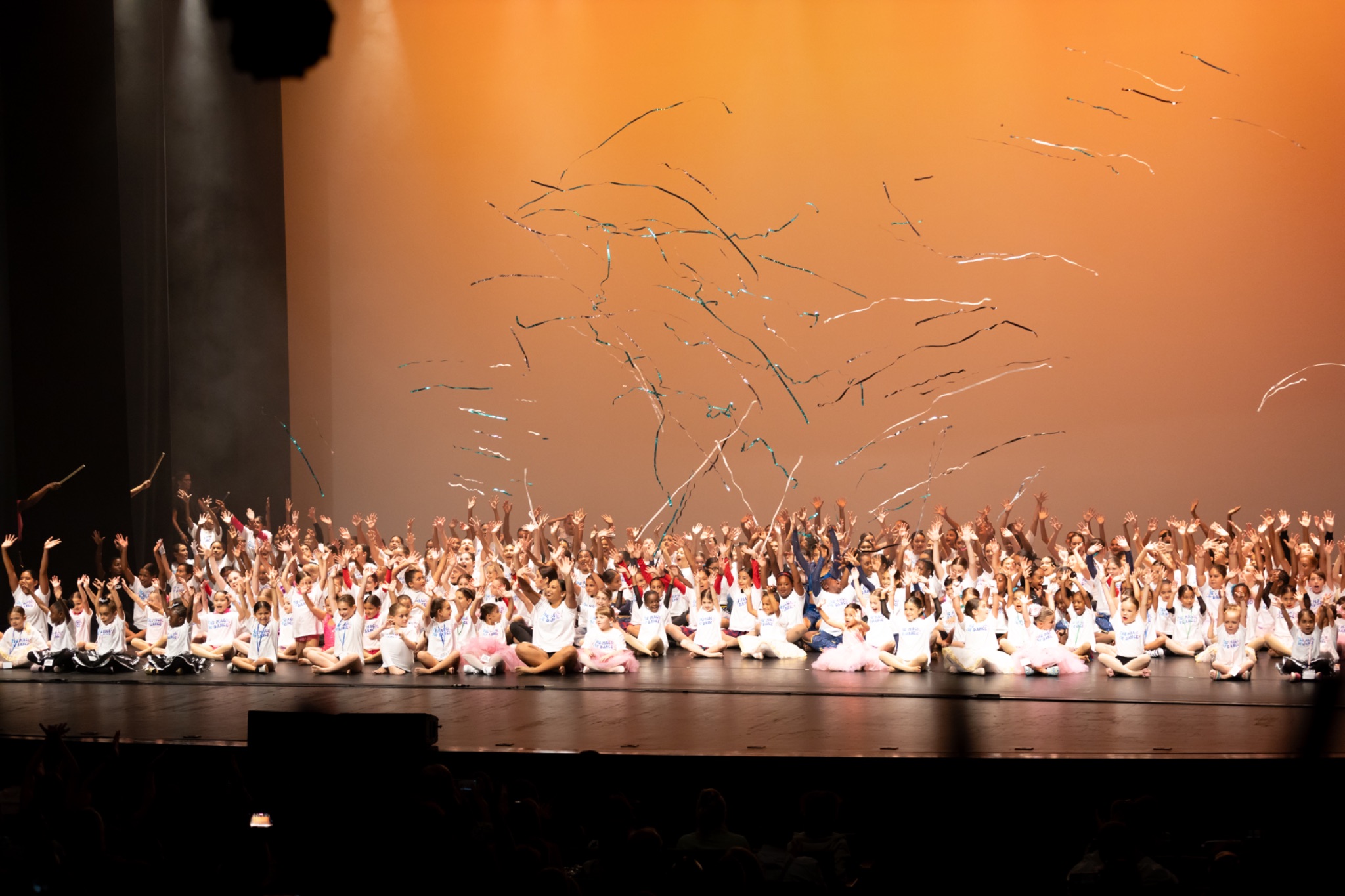 A group of dancers sits on the stage with their arms raised as silver confetti streamers fly over them.