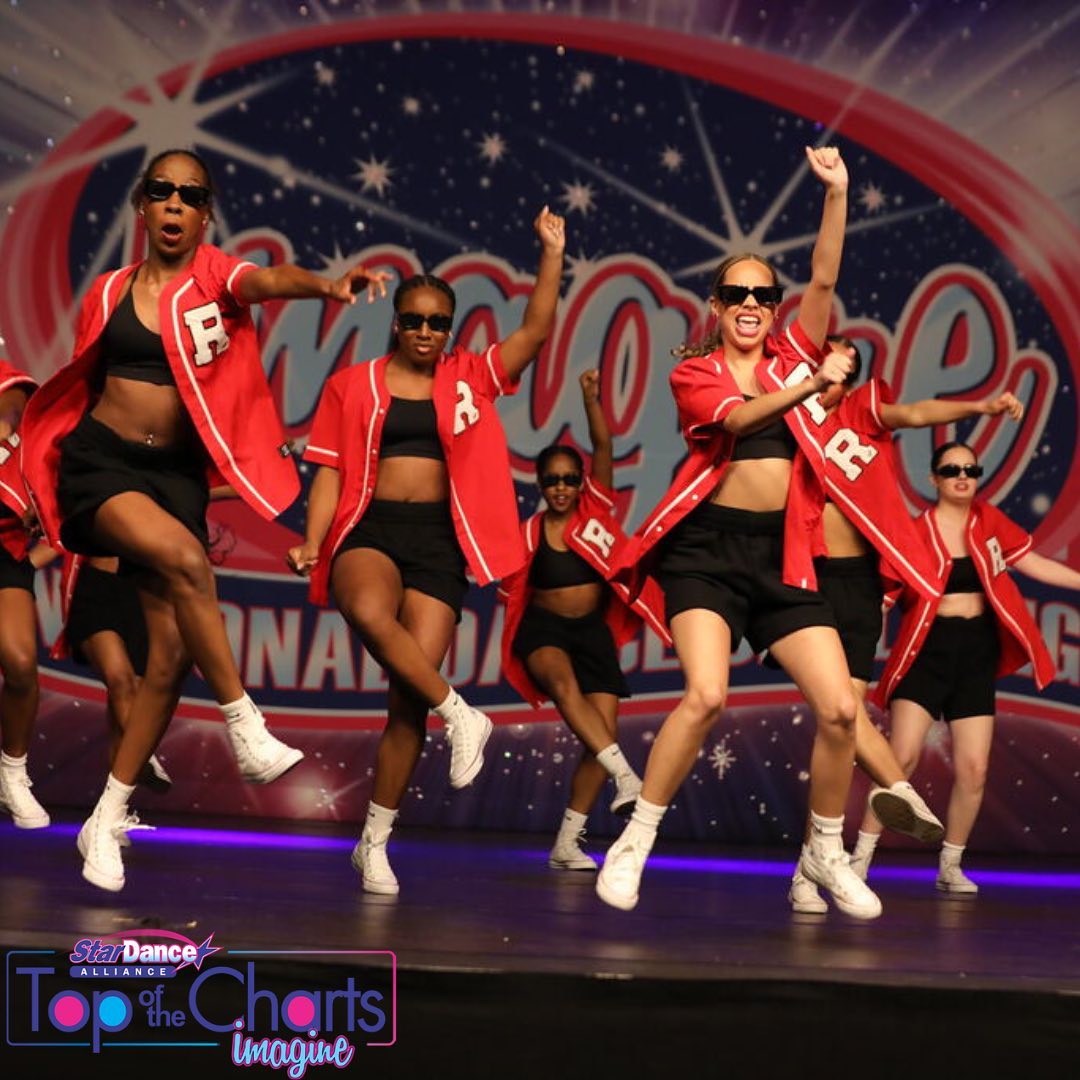Dancers mid-hop on stage wearing pink shirts, black shorts, and black sunglasses